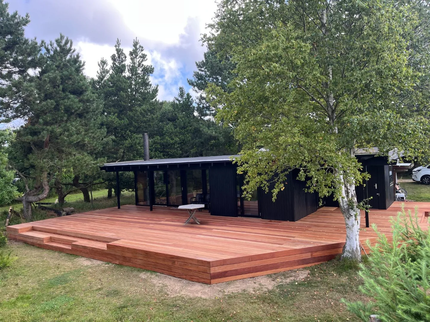 Jatoba-terrassen set på afstand, hele vejen rundt om et sort hus med fyrretræer og en birk i Liseleje.